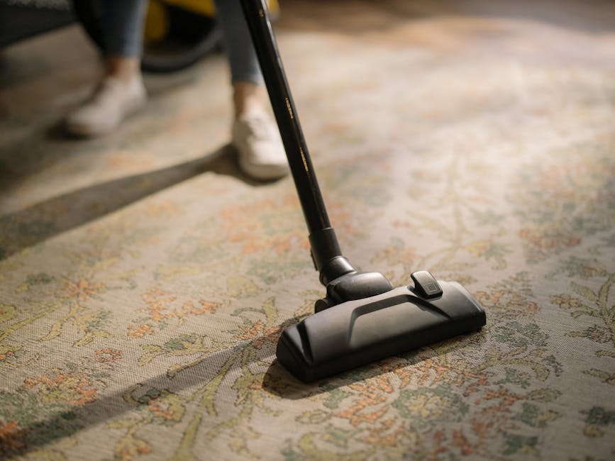 A woman with brown hair tied back, wearing a light blue striped shirt and jeans, is kneeling on a patterned rug in a living room, actively cleaning the wooden floor with a blue cloth. The room feature