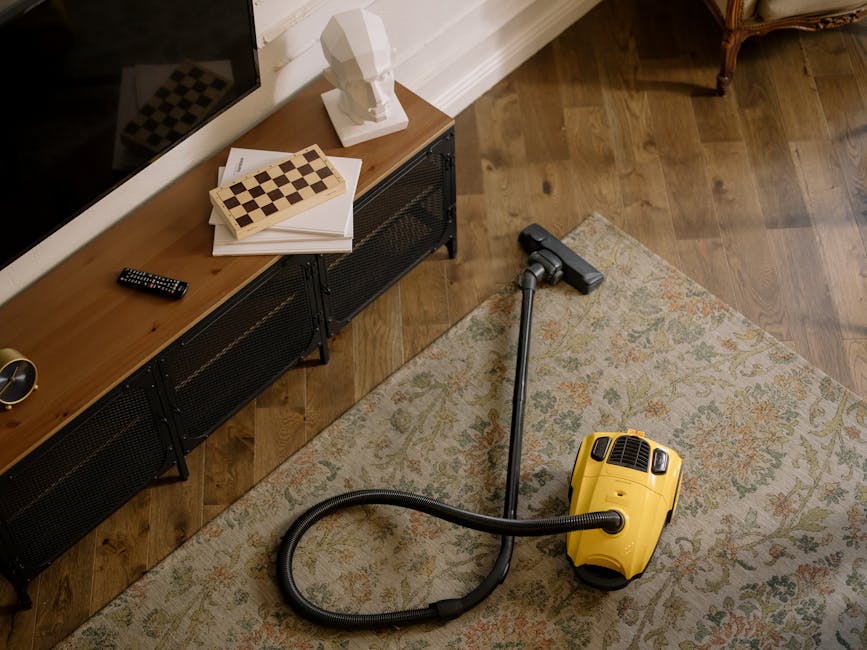A professional cleaner dressed in a white protective suit, face mask, and gloves is performing surface cleaning on a beige, plush carpet in a well-lit living room. The cleaner is using a long-handled