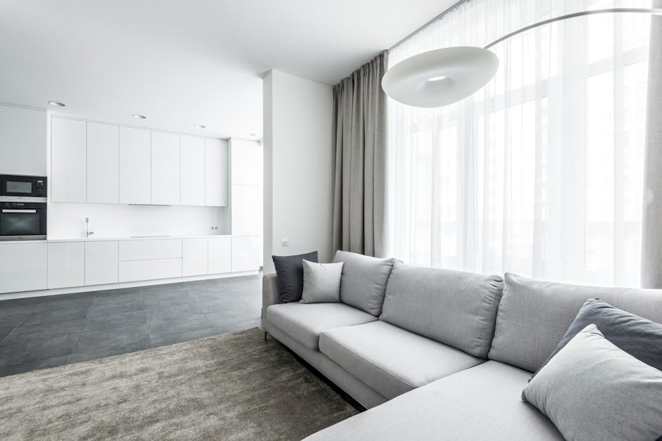A modern, minimalist living room featuring a light gray fabric sofa adorned with various gray cushions, positioned adjacent to large floor-to-ceiling windows with sheer white curtains allowing natural light to fill the space. The room has a neutral color palette with white walls and a beige area rug over a dark tiled floor. In the background, an open-plan kitchen with white cabinetry, built-in appliances, and a sleek countertop is visible. The overall scene depicts a clean, well-maintained domestic space with a bright, airy atmosphere, suitable for surface cleaning and domestic sanitisation services provided by Carpet Cleaning Finchley.