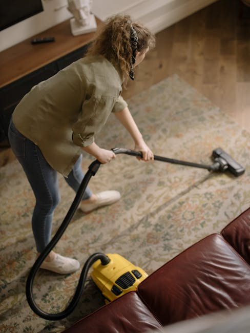 A woman with curly hair, dressed in a beige jacket and blue jeans, is engaged in surface cleaning within a living room. She is using a yellow vacuum cleaner with a black hose attachment to clean a patterned area rug that covers a wooden floor. The room features a brown leather sofa and a wooden sideboard on the side, with a remote control placed on top. The space is well-lit with natural light, highlighting the cleanliness and maintenance of the carpet and surrounding surfaces. This scene exemplifies domestic cleaning and deep cleaning practices as offered by Carpet Cleaning Finchley, captured in a typical North Finchley High Road home setting.